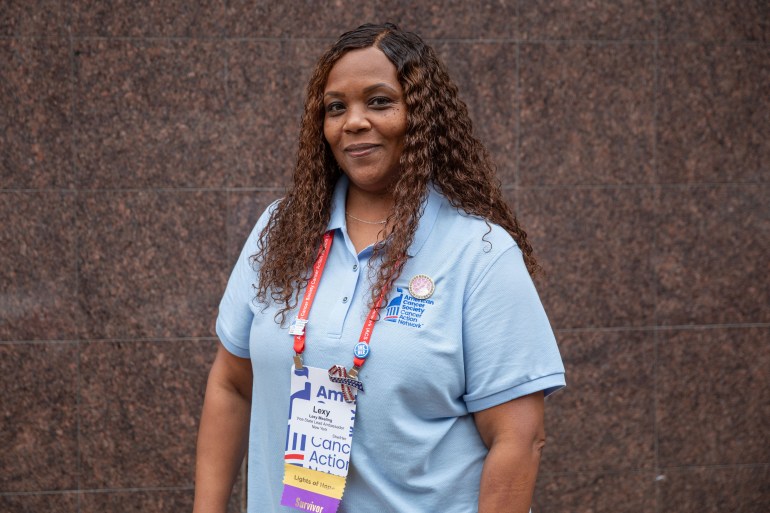 A portrait of a woman wearing a blue shirt standing against a brown background.