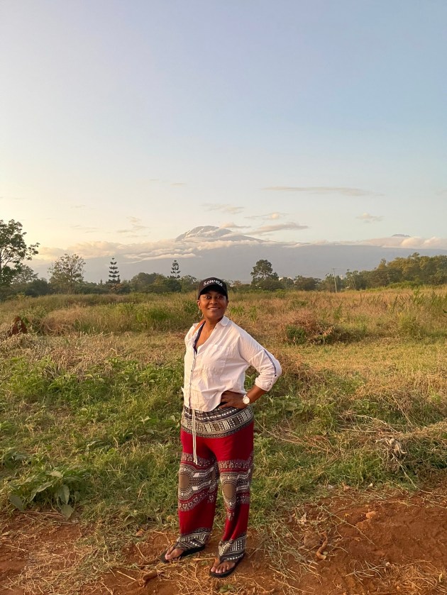 A woman stands in front of an open field with mountains in the distance behind her
