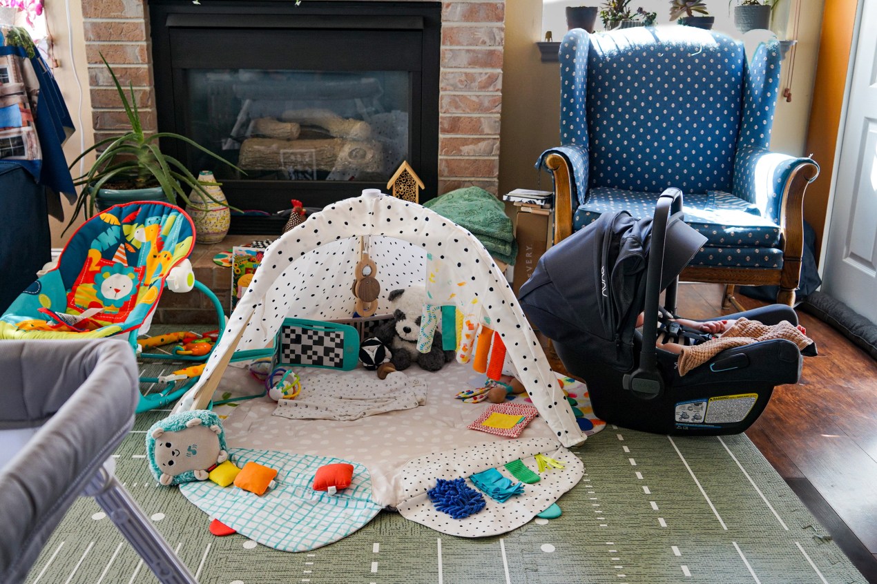 A photo of a baby in a carrier sleeping. Next to it is a play mat and a chair.