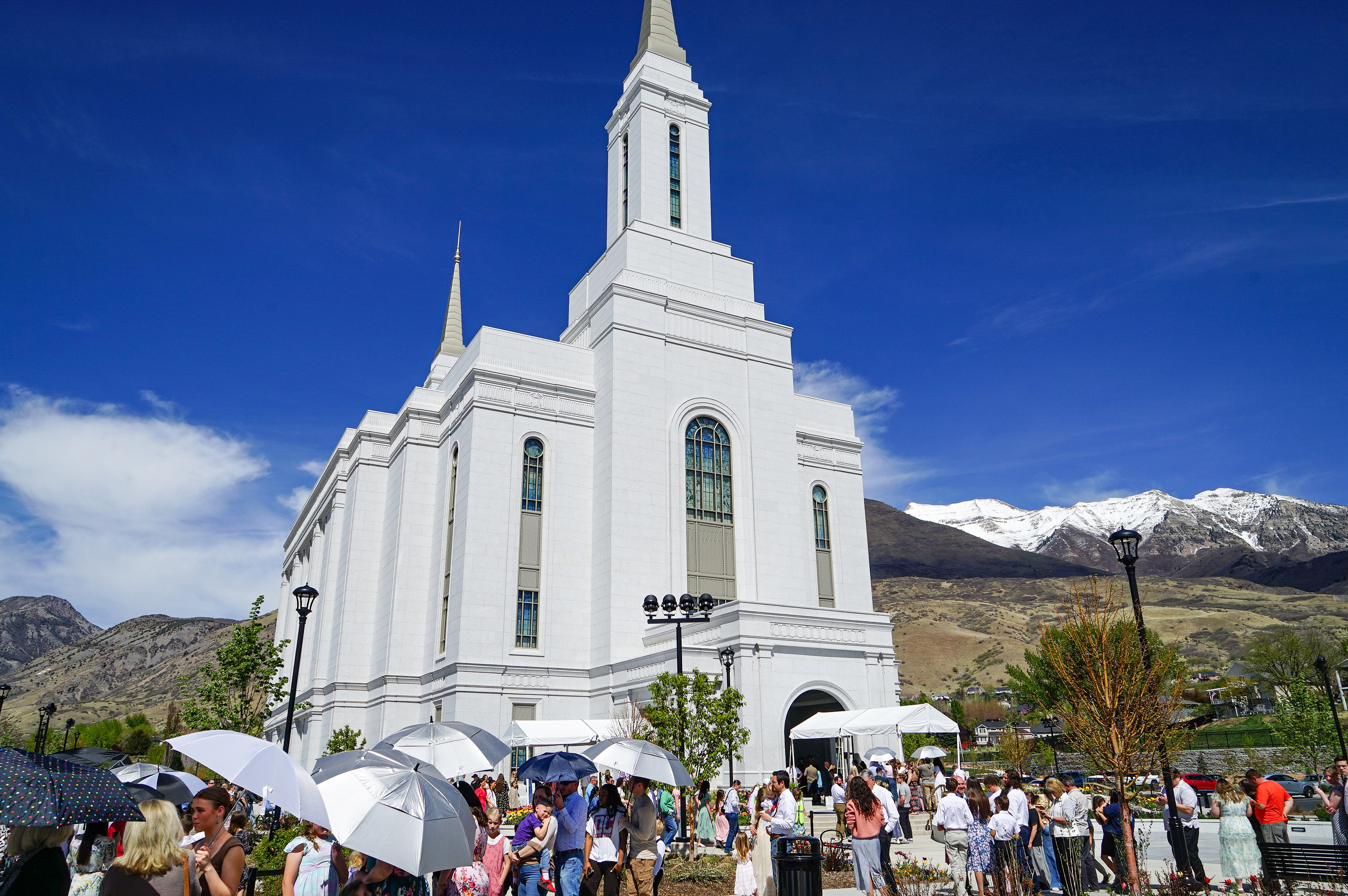 A photo of an LDS temple: a large white church. People are gathered in front of it, some of them holding umbrellas to protect from the sun.