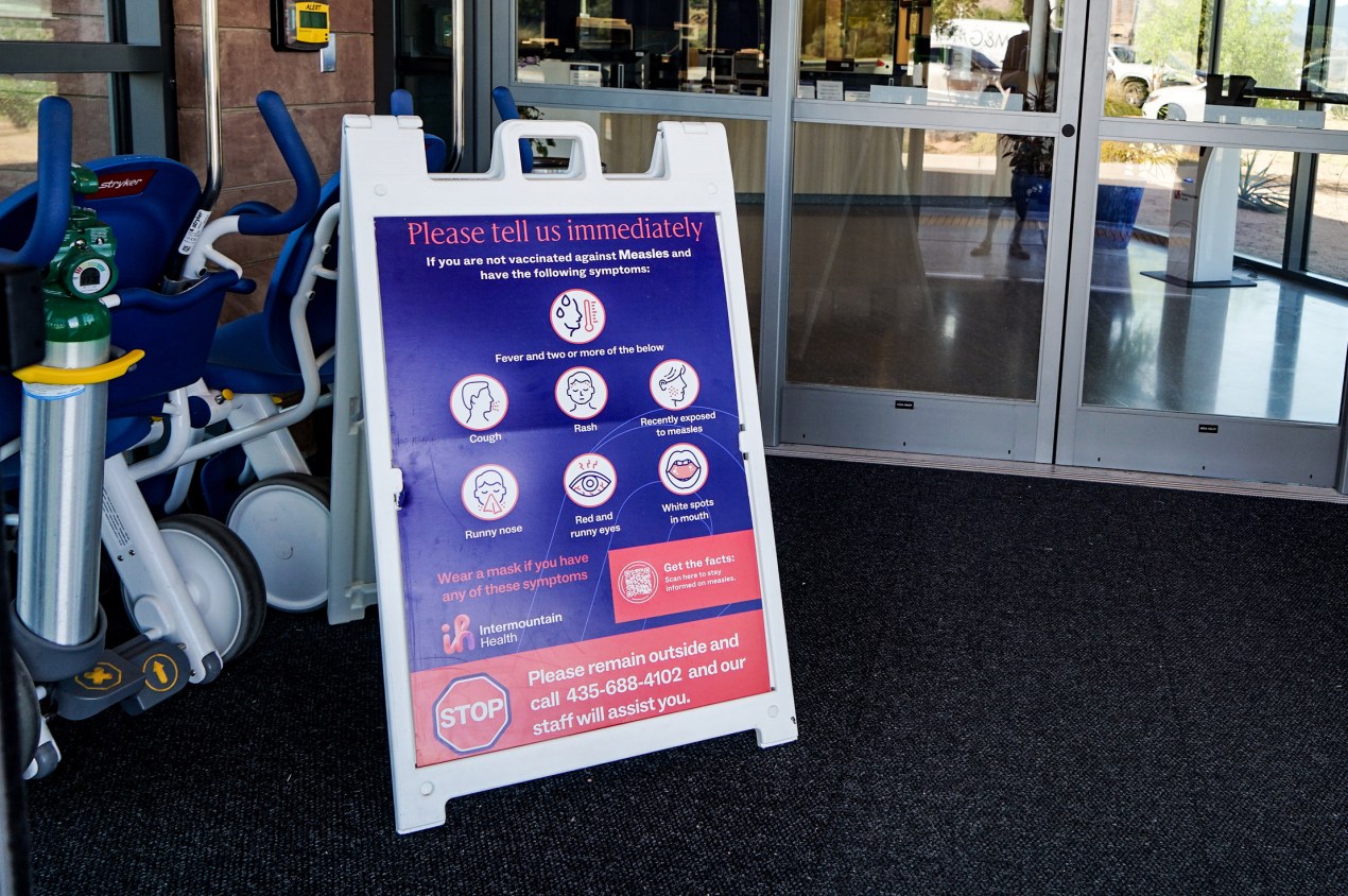 A sign in front of a hospital reads, "Please tell us immediately if you are not vaccinated against Measles and have the following symptoms: fever and two or more of the below — cough, rash, recently exposed to measles, runny nose, red and runny eyes, white spots in mouth."