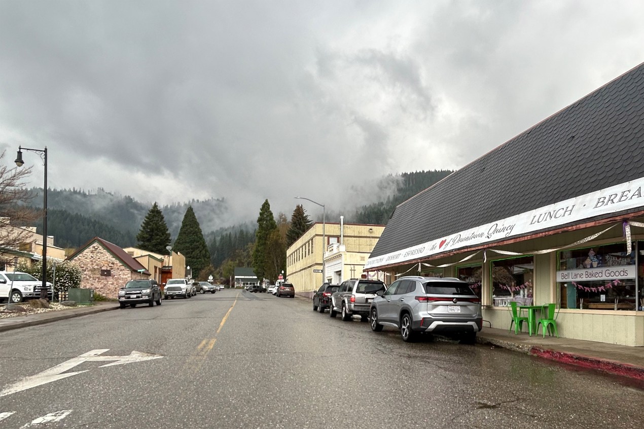A photo of a street lined with small shops in a Northern California mountain town. Fog rises over the treetops in the distance.