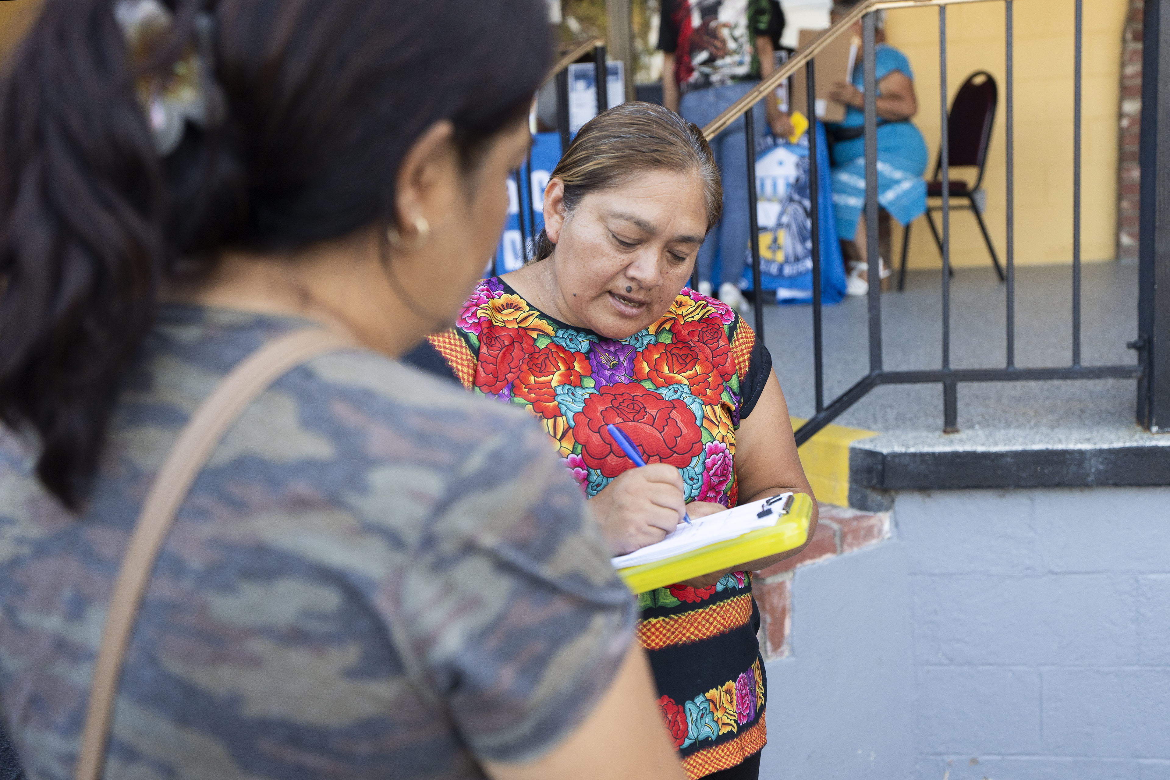 A woman writes on a clipboard as she speaks to another person whose back is to the camera.